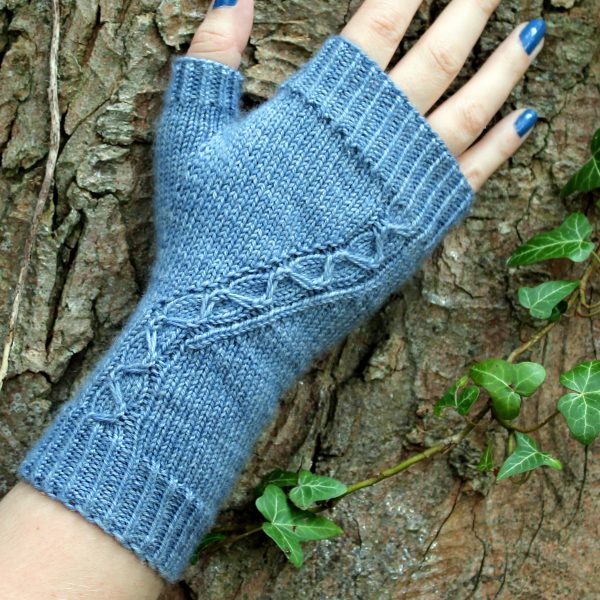 A blue fingerless mitt with a cable pattern travelling diagonally across the back of the hand, laid against an ivy covered tree