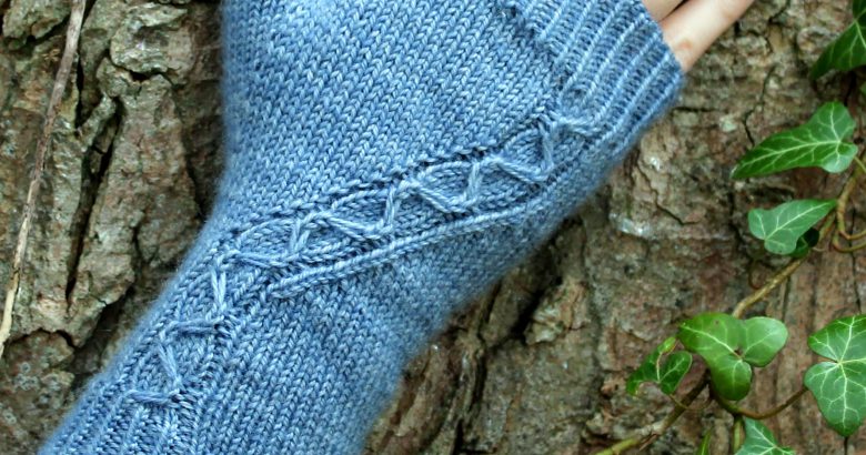 A blue fingerless mitt with a cable pattern travelling diagonally across the back of the hand, laid against an ivy covered tree