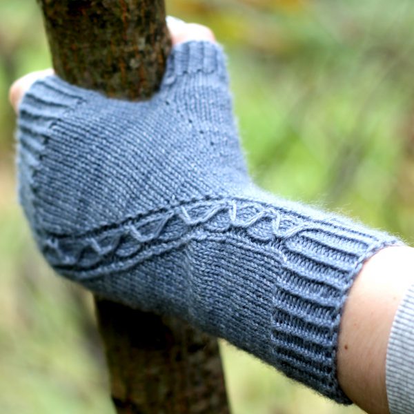 A blue fingerless mitt with a cable pattern travelling diagonally across the back of the hand, with the hand holding a tree branch