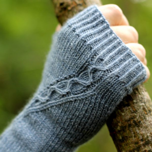 A blue fingerless mitt with a cable pattern travelling diagonally across the back of the hand, showing the detail of the cable meeting the cuff, with the hand holding a tree branch