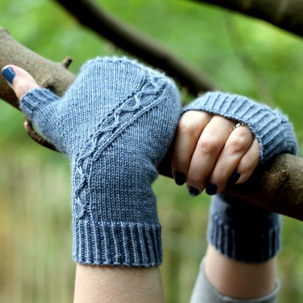 A blue fingerless mitt with a cable pattern travelling diagonally across the back of the hand, with both hands holding a tree branch