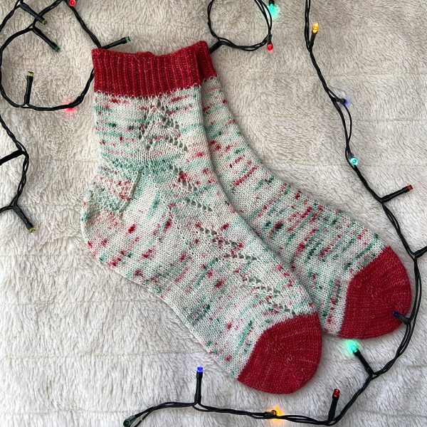 Giuthas socks knit in white yarn with red and green speckled and with red contrast toes and cuffs on a white rug surrounded by fairy lights