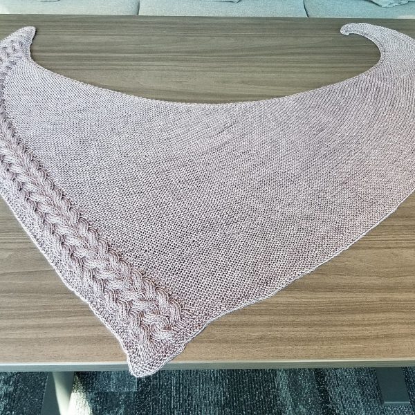 A large pale coloured asymmetrical garter stitch shawl with a reversible cable pattern down one edge laid on a table