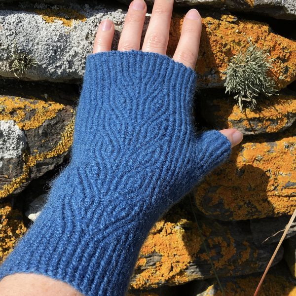 A dark blue fingerless mitt with twisted rib and a faux cable pattern, against a dry stone wall