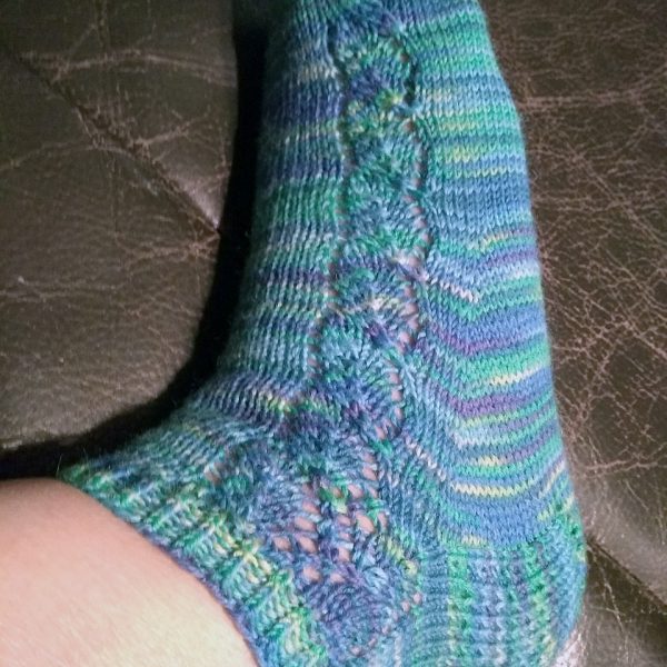 A sock with a lace leaf pattern up the outside of the foot knitted in variegated blue and purple yarn