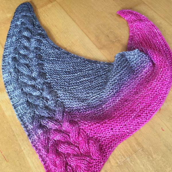 An asymmetrical garter stitch shawl with a reversible cable pattern down one edge with a grey to pink gradient