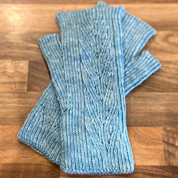 A pair of light blue fingerless mitts with twisted rib and a faux cable pattern, laid flat on a wooden surface