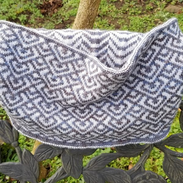 A cowl with stranded knitting in speckled grey and white yarn with a pattern made up of interlocking "V" shapes and scale shapes, over the back of a chair. You can see a colourwork pattern of soft curves on the inside