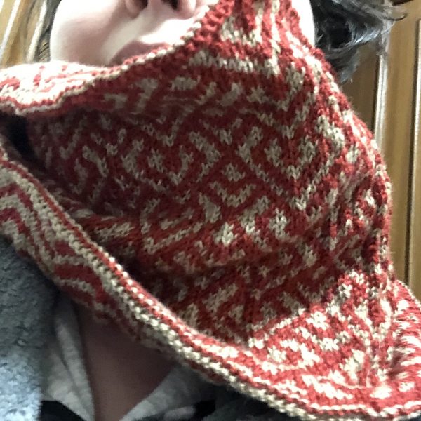 A cowl with stranded knitting in red and gold yarn with a pattern made up of interlocking "V" shapes and scale shapes, modelled by a woman