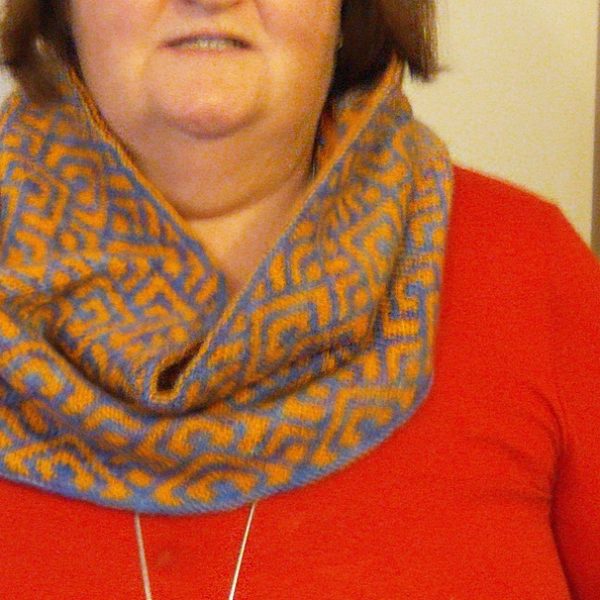 A cowl with stranded knitting in orange and blue yarn with a pattern made up of interlocking "V" shapes and scale shapes, modelled woman in a red top