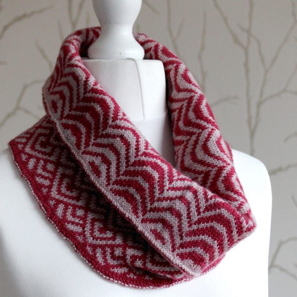 A cowl with stranded knitting in red and pink yarn with a pattern made up of soft curves, folded over a pattern made up of interlocking "V" shapes and scale shapes, modelled on a mannequin