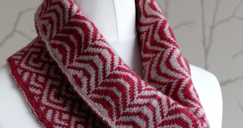 A cowl with stranded knitting in red and pink yarn with a pattern made up of soft curves, folded over a pattern made up of interlocking "V" shapes and scale shapes, modelled on a mannequin