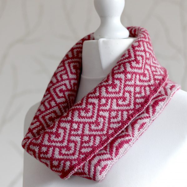 A cowl with stranded knitting in red and pink yarn with a pattern made up of interlocking "V" shapes and scale shapes, folded over a pattern made up of soft curves, modelled on a mannequin