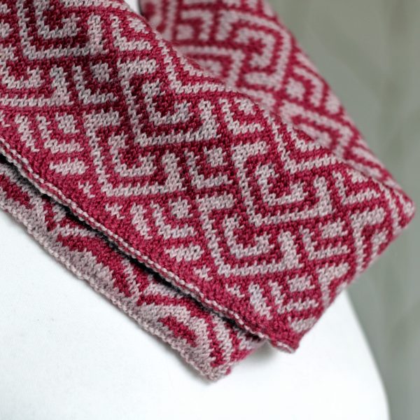 A close up of a cowl with stranded knitting in red and pink yarn with a pattern made up of interlocking "V" shapes and scale shapes, folded over a pattern made up of soft curves, modelled on a mannequin