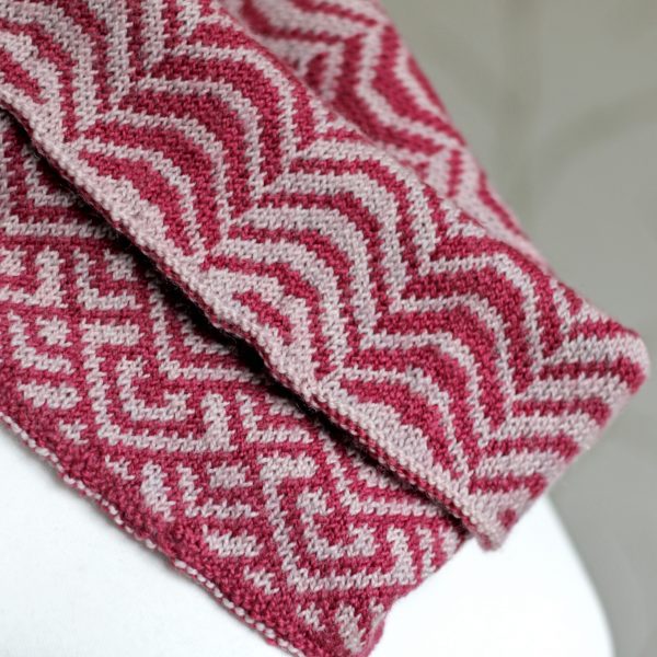 A close up of a cowl with stranded knitting in red and pink yarn with a pattern made up of soft curves, folded over a pattern made up of interlocking "V" shapes and scale shapes, modelled on a mannequin
