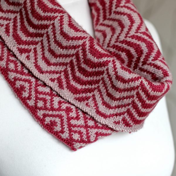 A close up of a cowl with stranded knitting in red and pink yarn with a pattern made up of soft curves, folded over a pattern made up of interlocking "V" shapes and scale shapes, modelled on a mannequin