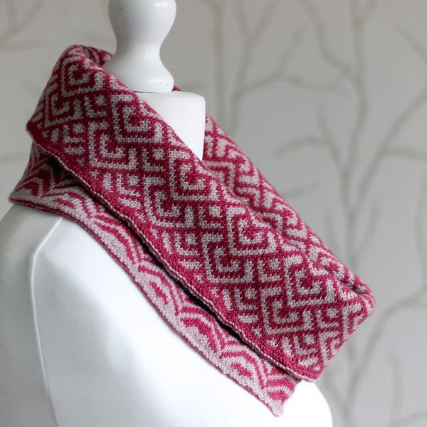 A cowl with stranded knitting in red and pink yarn with a pattern made up of interlocking "V" shapes and scale shapes, folded over a pattern made up of soft curves, modelled on a mannequin