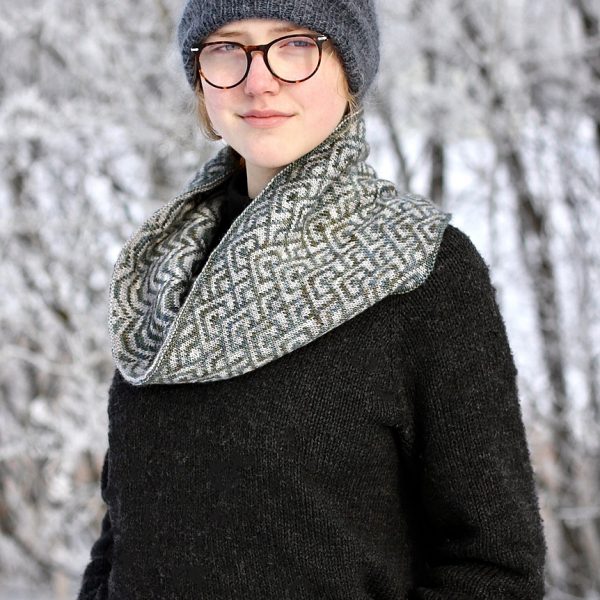 A cowl with stranded knitting in grey and white speckled yarn with a pattern made up of interlocking "V" shapes and scale shapes, modelled by a girl wearing glasses