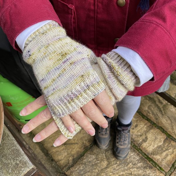 Two hands wearing a pair fingerless mitts knit in yellow and grey yarn with colourful strips showing the garter stitch columns around the thumb gusset and the outside of the hand