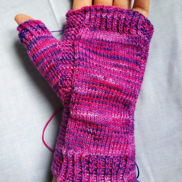 A hand wearing a fingerless mitt knit in variegated yarn showing the garter stitch columns around the thumb gusset and the outside of the hand