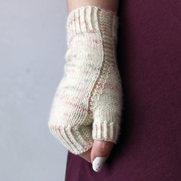 A hand making a fist showing the garter stitch column wrapping around the thumb gusset of a fingerless mitt knit in cream yarn with pink and green speckles