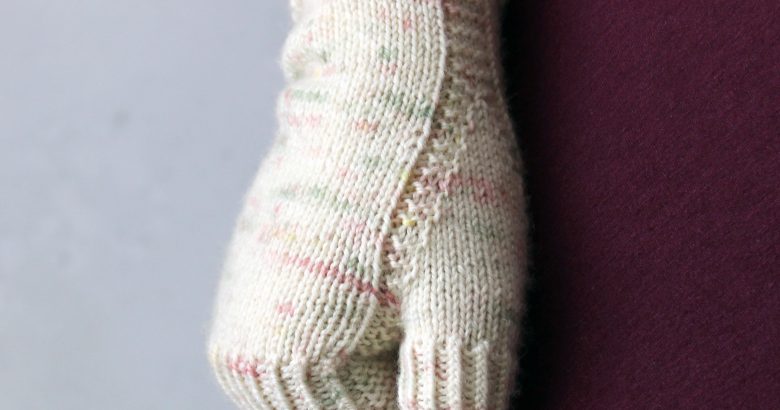 A hand making a fist showing the garter stitch column wrapping around the thumb gusset of a fingerless mitt knit in cream yarn with pink and green speckles