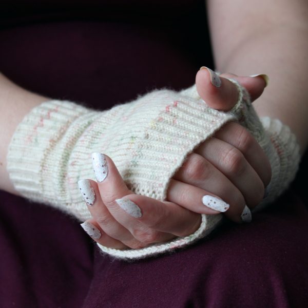 Two hands clasped showing the garter stitch column around the outside of one hand and the twisted rib cuff of a pair of fingerless mitts knit in cream yarn with pink and green speckles