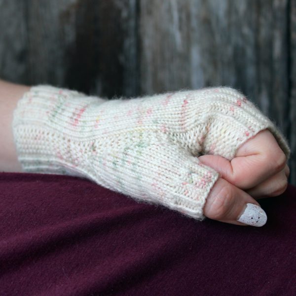 A hand making a fist showing the garter stitch column wrapping around the thumb gusset of a fingerless mitt knit in cream yarn with pink and green speckles