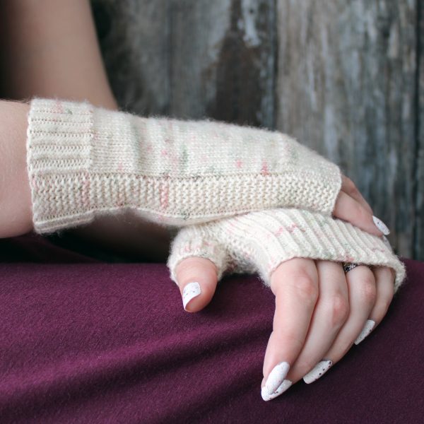 Two hands clasped showing the garter stitch column around the outside of one hand and the column wrapping around the thumb gusset of the other hand of a pair of fingerless mitts knit in cream yarn with pink and green speckles