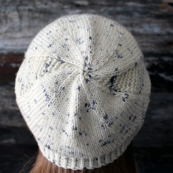 The crown of a slouchy hat knit in white yarn with black speckles and garter stitch columns up each side