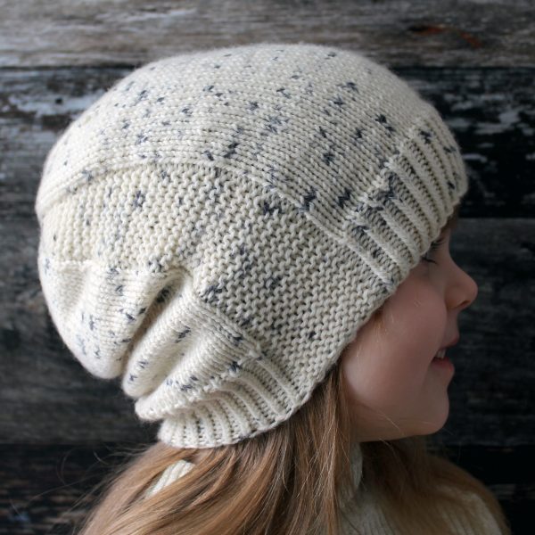 A young girl wearing a slouchy hat knit in white yarn with black speckles and garter stitch columns up each side