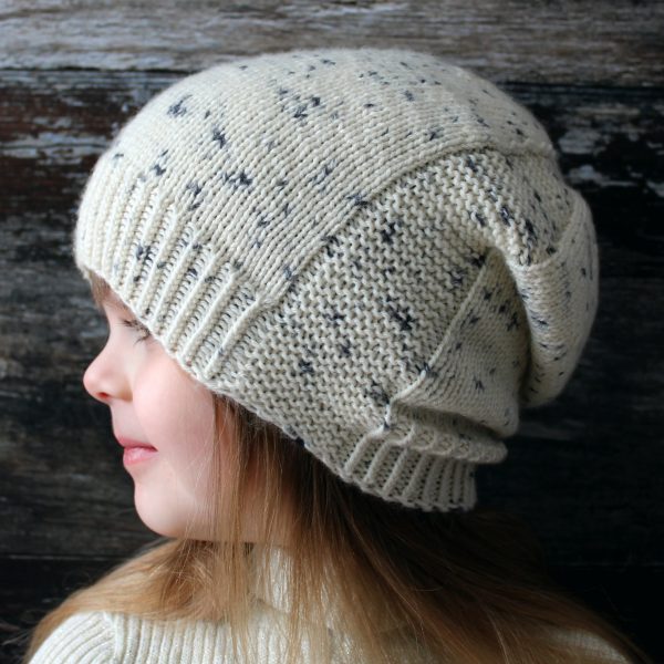 A young girl wearing a slouchy hat knit in white yarn with black speckles and garter stitch columns up each side