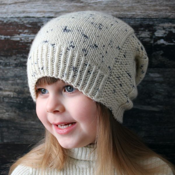 A young girl wearing a slouchy hat knit in white yarn with black speckles and garter stitch columns up each side