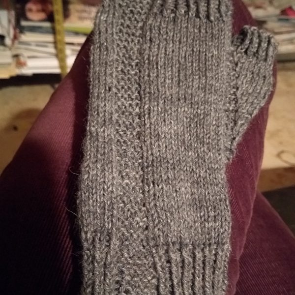 A fingerless mitt knit in grey yarn showing the garter stitch columns around the thumb gusset and the outside of the hand