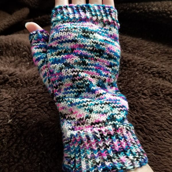 A hand wearing a fingerless mitt knit in variegated yarn with colourful strips showing the garter stitch columns around the thumb gusset and the outside of the hand