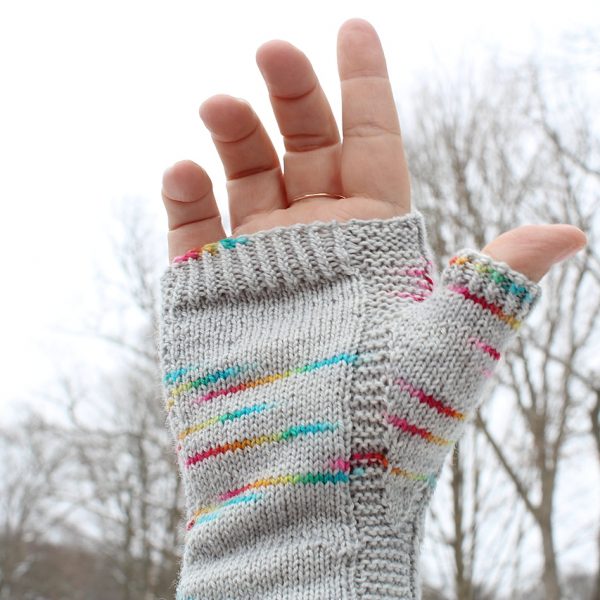 A hand wearing a fingerless mitt knit in grey yarn with colourful strips showing the garter stitch columns around the thumb gusset and the outside of the hand