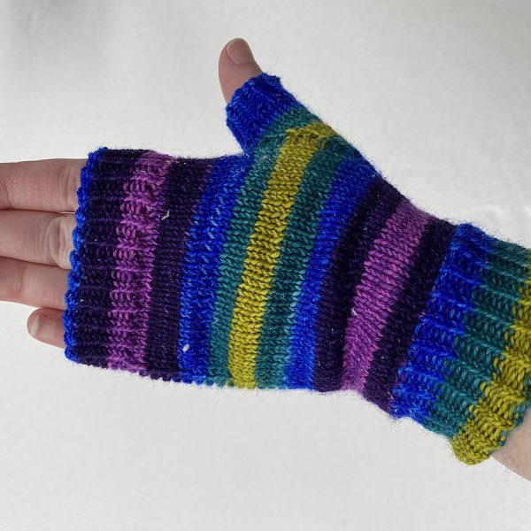 A hand wearing a fingerless mitt knit in striped yarn with colourful strips showing the garter stitch columns around the thumb gusset and the outside of the hand