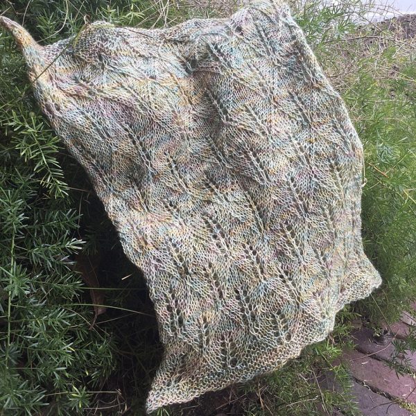 A pale variegated coloured cowl with diamond shaped lace leaves and wide garter stitch borders draped over a bush