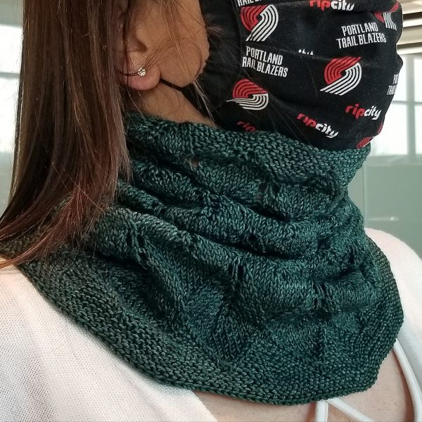 A model wearing a cowl in dark green yarn with diamond shaped lace leaves and a wide garter stitch border