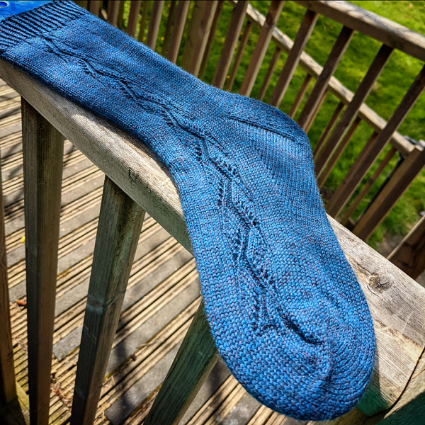 A sock handknit in blue yarn laid flat to show the zigzag lace pattern up the outside of the foot and leg