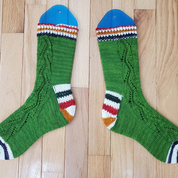 A pair of socks handknit in green yarn with contrast heels and toes laid flat to show the zigzag lace pattern up the outside of each foot and leg