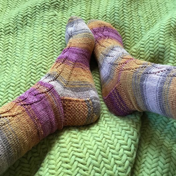 A pair of socks handknit in striped yarn showing the zigzag lace pattern up the outside of one foot and leg,