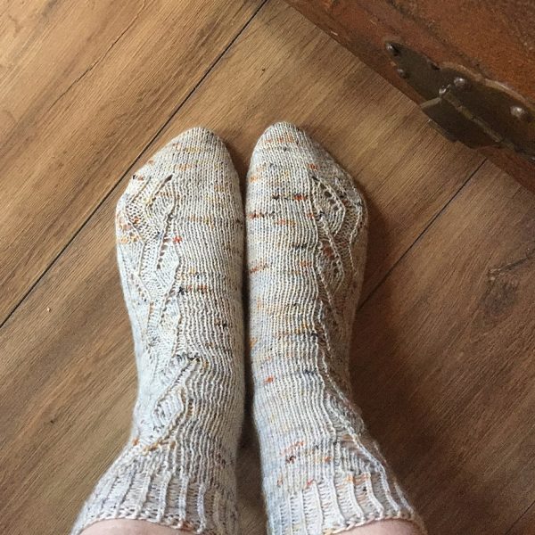 A pair of socks handknit in white speckled yarn showing the zigzag lace pattern up the outside of each foot and leg,