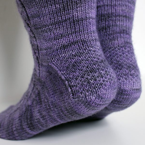 A pair of socks handknit in purple yarn showing textured heel pattern on both socks