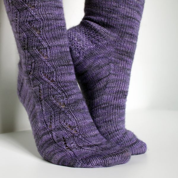 A pair of socks handknit in purple yarn showing the zigzag lace pattern up the outside of one foot and leg, and showing the gusset and textured heel pattern on the other foot