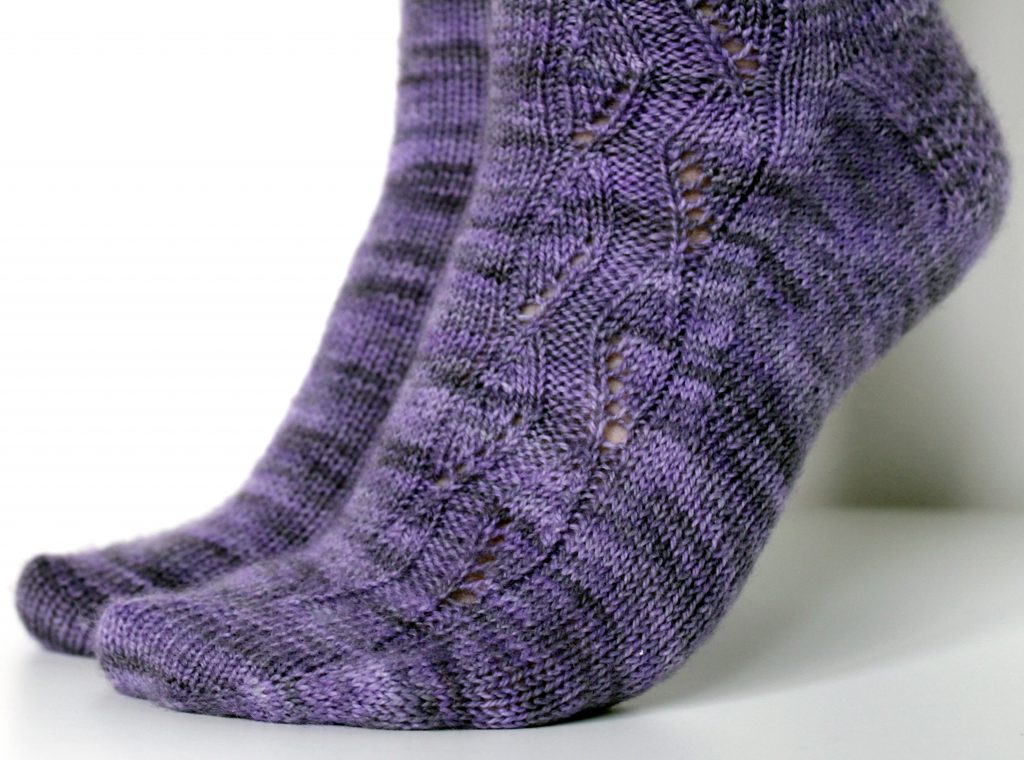 A pair of socks handknit in purple yarn showing the zigzag lace pattern up the outside of one foot and leg,