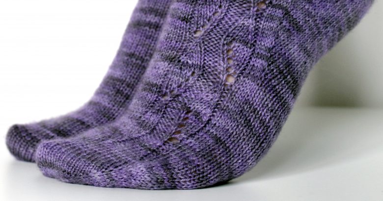 A pair of socks handknit in purple yarn showing the zigzag lace pattern up the outside of one foot and leg,