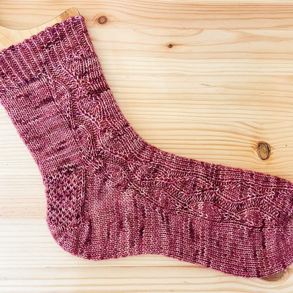 A sock handknit in pink yarn laid flat to show the zigzag lace pattern up the outside of the foot and leg