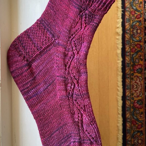 A sock handknit in red/purple yarn on a sock blocker to show the zigzag lace pattern up the outside of the foot and leg