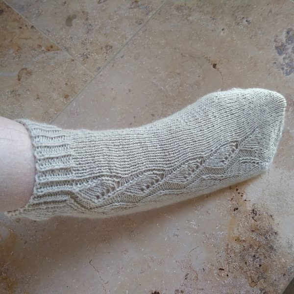 A sock handknit in white yarn showing the zigzag lace pattern up the outside of the foot and leg
